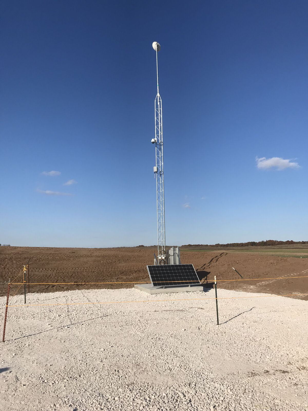 Solar-powered communications tower with a dish and security camera on a prepared gravel pad beside a ploughed field